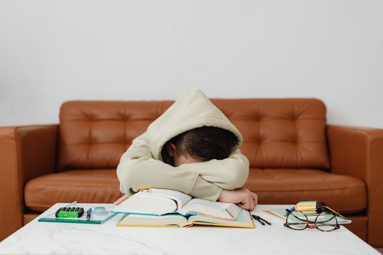 child in beige hoodie leaning forward on table feeling exhausted while studying